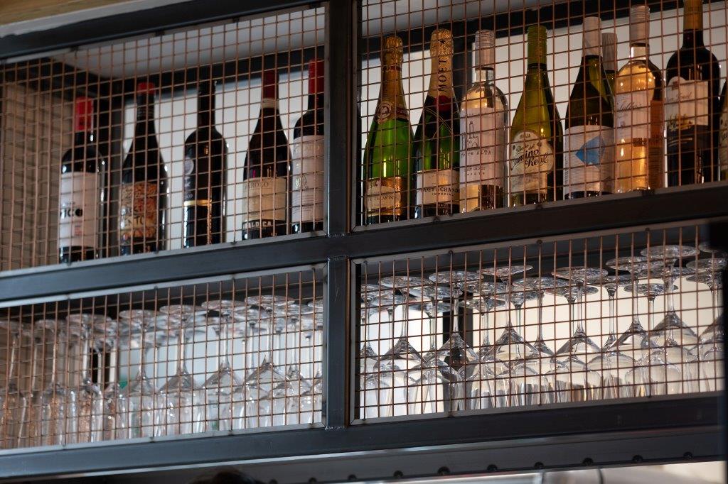 Bottles of wine and champagne are displayed on a top shelf behind a wire mesh, with rows of upside-down wine glasses hanging below on a glass rack.