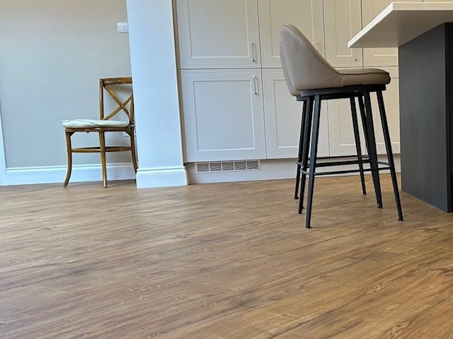 A kitchen with wood-look flooring, white cabinets, a beige barstool at a dark island, and a wooden chair with a cushion against the wall in the background.