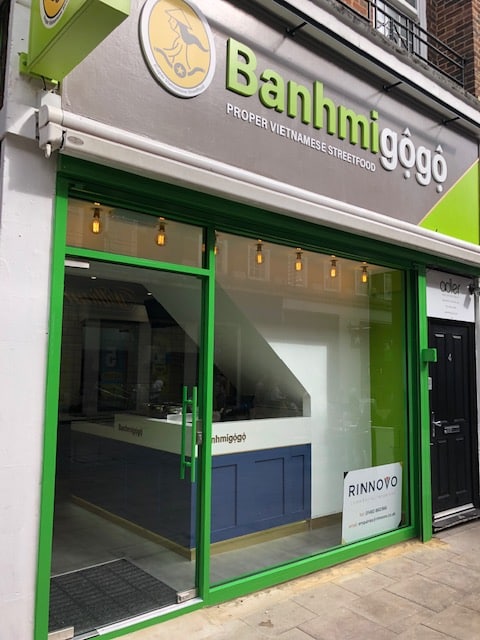 A Vietnamese street food restaurant named Banhmigogo with a gray and green exterior, large glass windows, and a modern, inviting look. A sign above the door reads Proper Vietnamese Streetfood.