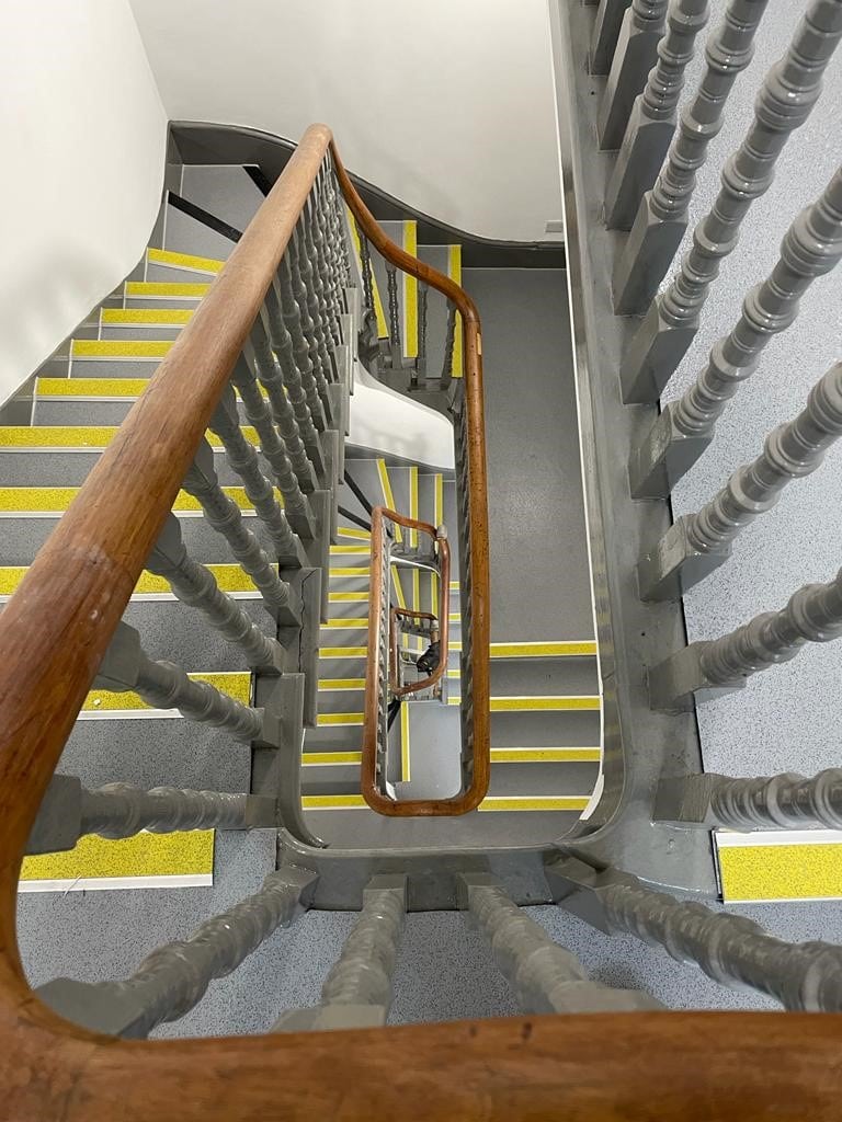 A spiral staircase with grey steps, yellow anti-slip strips, and light grey railings, viewed from above, creating a geometric and repetitive pattern down several floors.