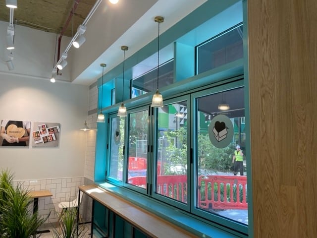 Bright café interior with large blue-framed windows, hanging pendant lights, wooden counters, and white tiled walls. Outside, a construction worker and red barricades are visible through the glass.