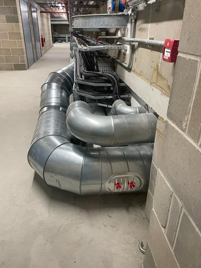 Large silver ventilation ducts with red valve handles run along the floor of an industrial hallway with concrete block walls and exposed wiring overhead.