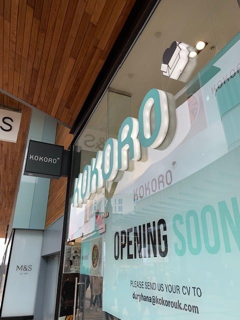 The photo shows the exterior of a KOKORO restaurant with large letters and a sign reading OPENING SOON. An email address for submitting CVs is displayed on the glass window. M&S store signage is visible nearby.