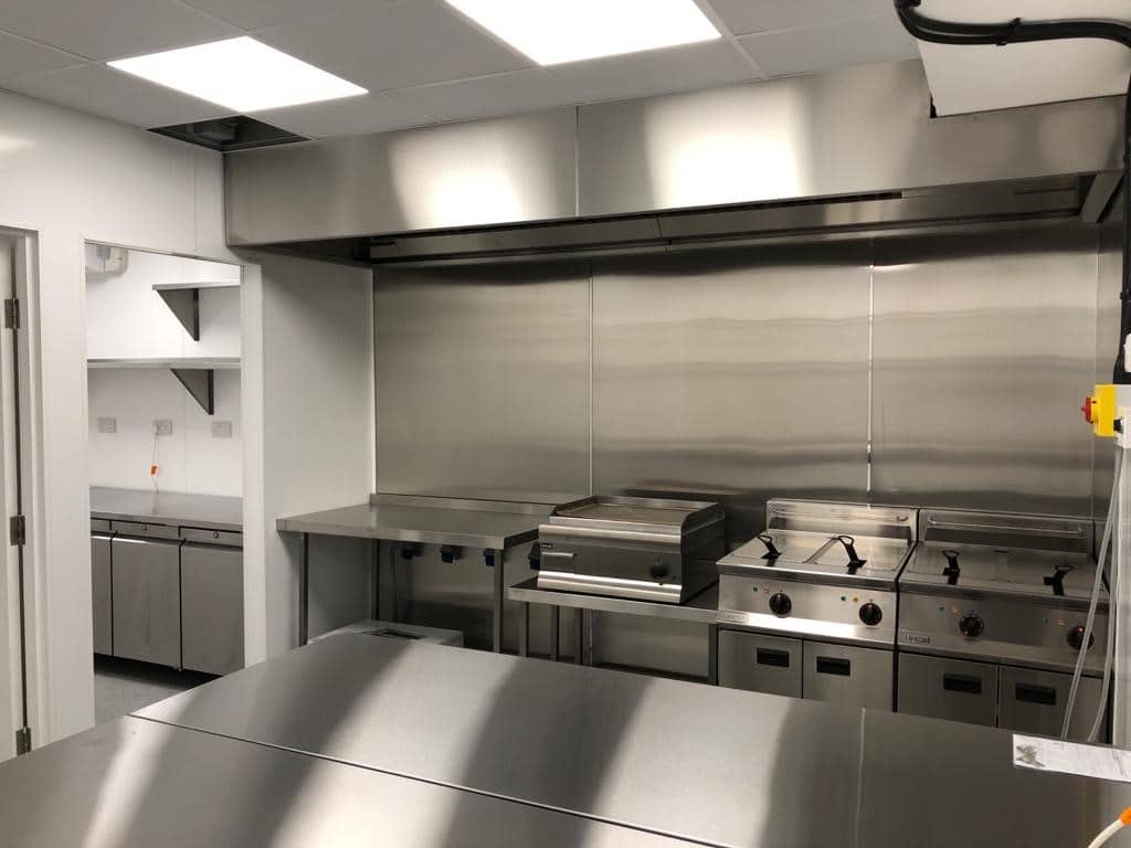 A commercial kitchen with stainless steel appliances, including grills and stoves, under a large ventilation hood. The counters and walls are also stainless steel, and there is a preparation area in the background.