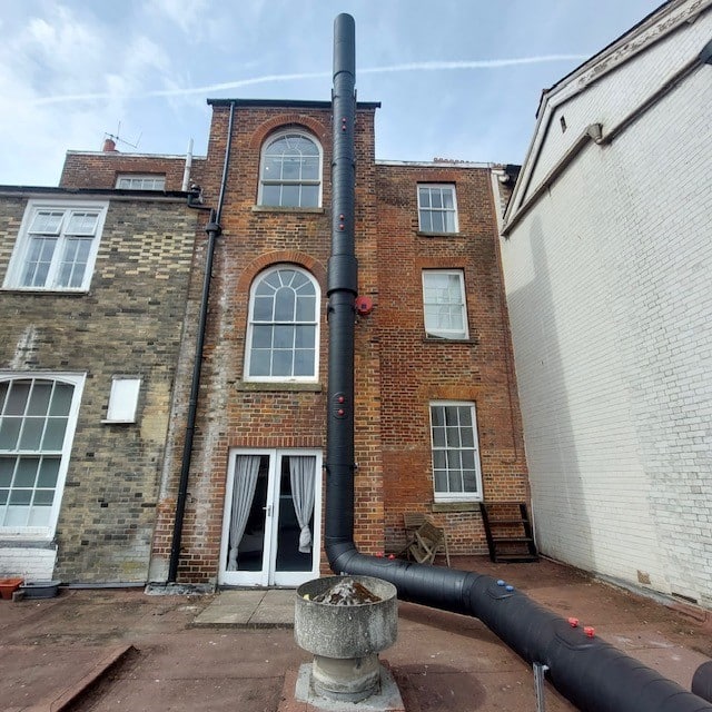A tall black pipe runs vertically from a stone planter on the ground up the exterior of a three-story brick building, passing by arched and rectangular windows. The pipe is supported with red brackets.