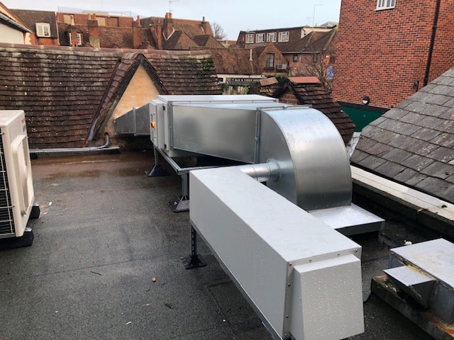 A rooftop view showing large silver HVAC ductwork and air conditioning units, surrounded by sloped tiled roofs and brick buildings in the background.