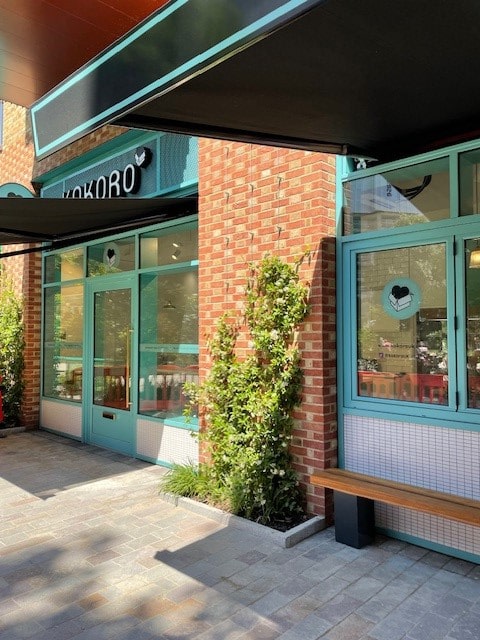 A brick storefront with turquoise window frames and door, a sign reading KOKORO, a wooden bench, green plants climbing the facade, and striped awnings shading the entrance on a sunny day.