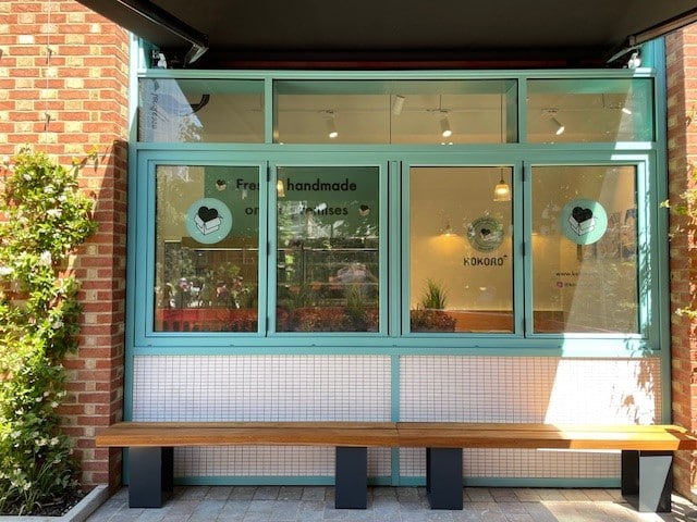 About Rinnovo: A storefront with large turquoise-framed windows and a long wooden bench outside. The window displays text: Fresh handmade onigiri & katsu houses, heart icons, and the KOKORO logo. Brick walls and plants surround the area.