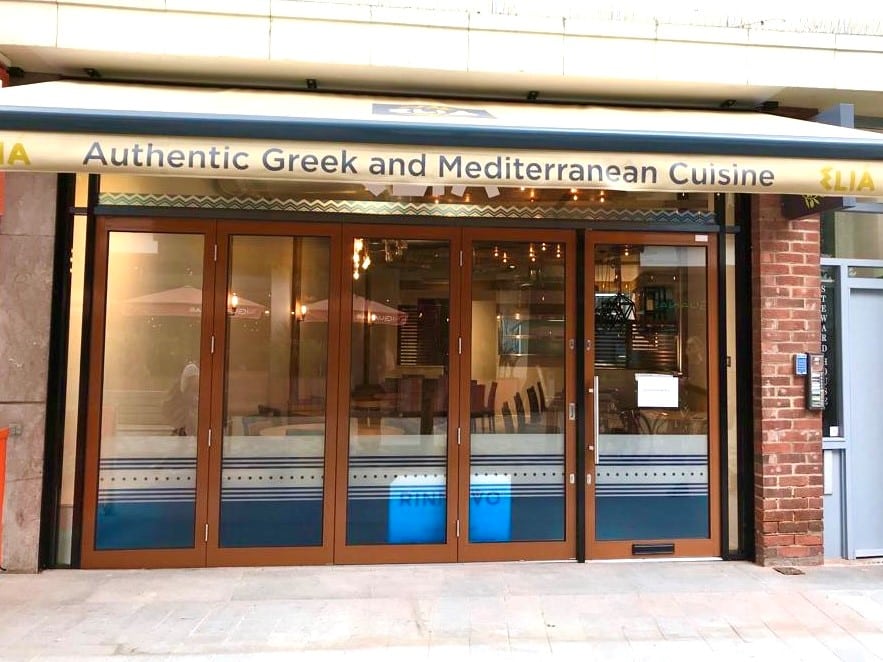 The exterior of a restaurant features large glass doors, Awnings and Cafe Barriers, and a sign reading Authentic Greek and Mediterranean Cuisine. The lights inside are on, with a small notice posted on one of the doors.