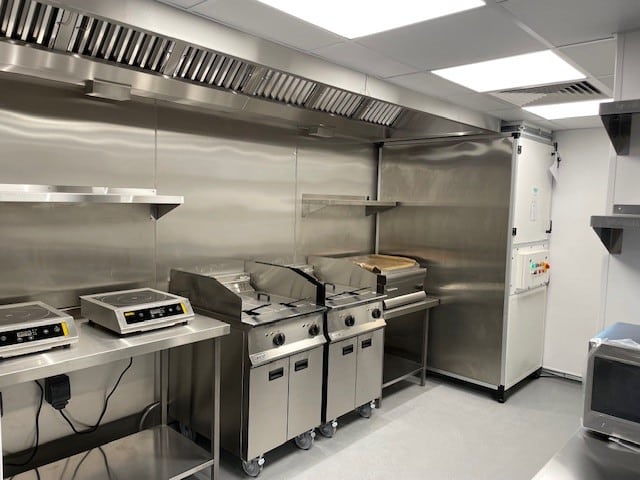A clean, modern commercial kitchen with stainless steel surfaces, fryers, induction cooktops, ventilation hood, shelves, and control panels, all neatly arranged against the walls.