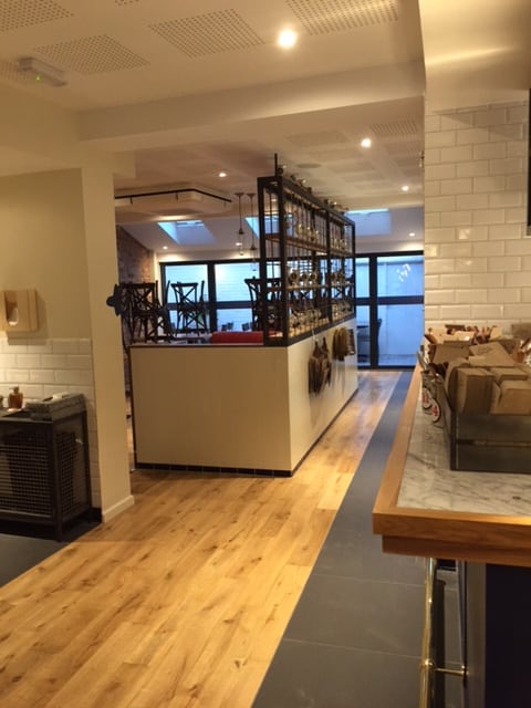 A modern restaurant interior with wooden flooring, tiled walls, and upside-down chairs stacked on tables. The space is empty, warmly lit, and clean, with a counter on the right and glass doors at the back.