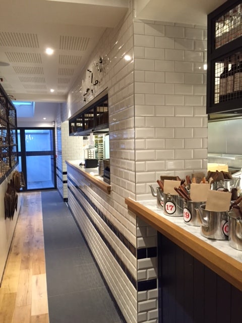 A narrow, modern restaurant interior with white subway tile walls, wood flooring, a counter with metal buckets holding utensils, and a view toward the entrance with natural light coming in.