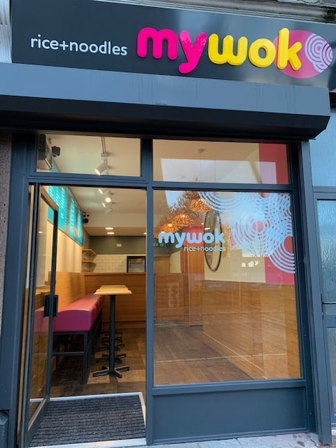 Storefront of mywok restaurant with large glass windows, a visible logo, and a modern interior featuring wooden tables, high stools, and a bright pink cushioned bench. The sign above reads rice+noodles mywok in bold letters.