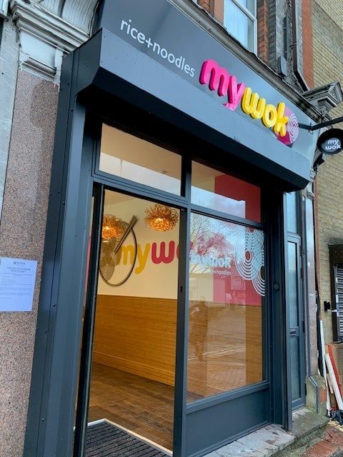 Storefront of a restaurant called my wok, with colorful sign text. Reflections of trees and hanging lights are visible in the glass windows; the words rice+noodles appear beside the restaurant’s name on the black awning.