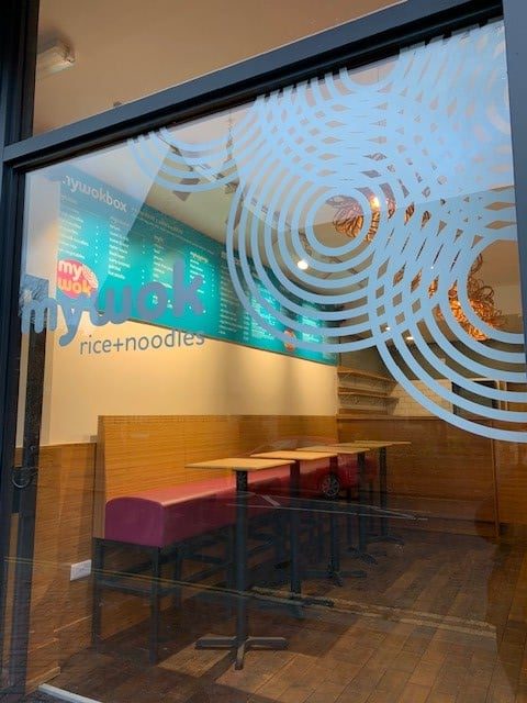 View through a window decorated with white circular patterns into a restaurant with wooden floors, pink seating, and a blue menu board on the wall labeled my wok rice+noodles. Tables and chairs are stacked.