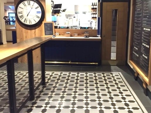 A restaurant counter area with a patterned black-and-white tile floor, a large wall clock, a wooden counter with metal mesh sides, and a menu board on the wall next to a door with a narrow window.