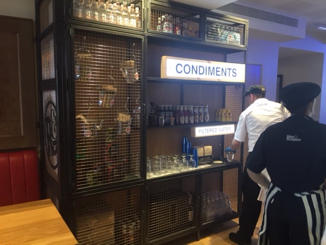 A condiment and water station in a restaurant features labeled shelves with sauces, glasses, and bottles. Two staff members in uniforms are nearby, one facing the counter and the other walking away.