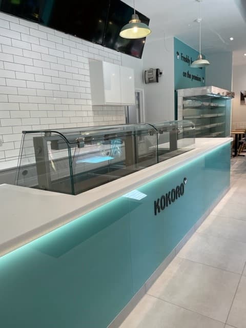 A clean, modern counter inside a Kokoro restaurant, featuring glass partitions, white tile walls, and a light blue front panel with the Kokoro logo. Empty food display units and shelves are visible.