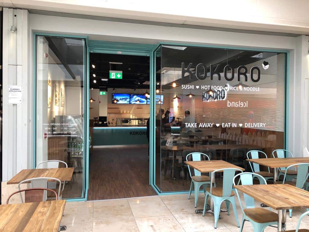 Street view of Kokoro restaurant with large glass windows, tables and chairs outside, and signage advertising sushi, hot food, rice, noodles, takeaway, eat-in, and delivery options. The interior is visible through open doors.