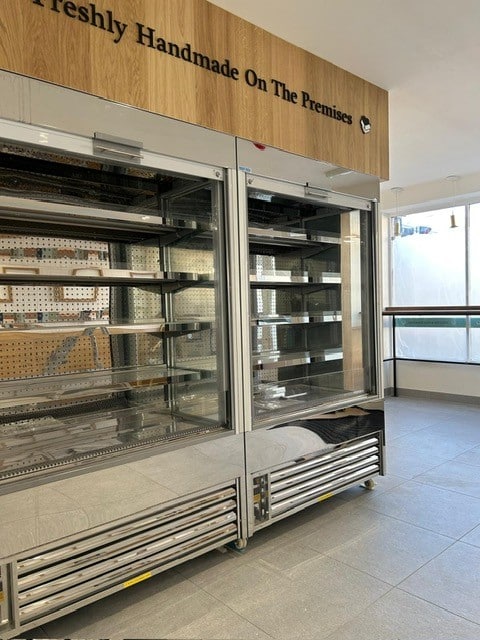 Empty commercial refrigerator display cases with glass doors and metal shelves in a clean, well-lit store. A sign above reads Freshly Handmade On The Premises. Sunlight streams through large windows.